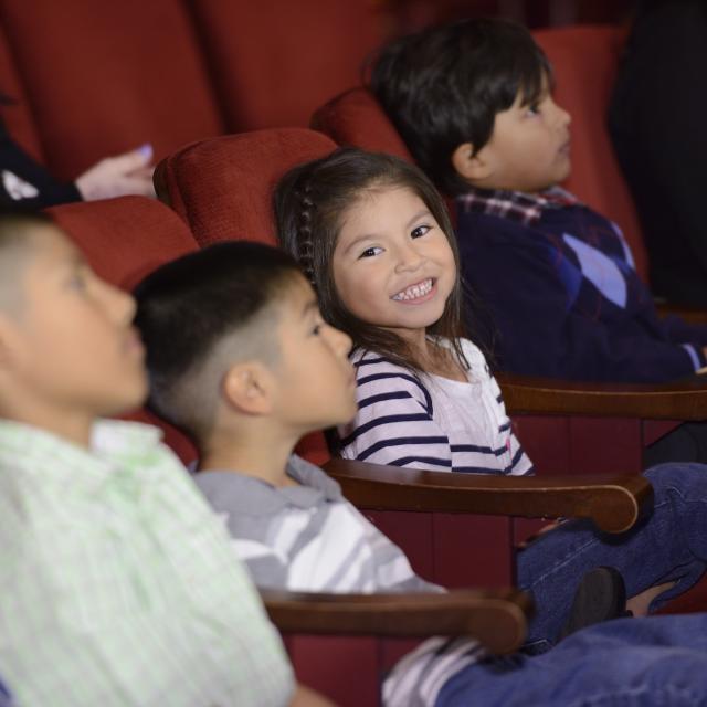 Classics 4 Kids A young girl, seated in a row with other students at a theatre, smiles at the camera.