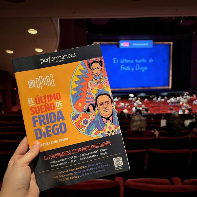 A hand holds up a paper program that reads "El Ultimo Sueno de Frida y Diego." Behind the program is a stage reading the same, with a blue background curtain.